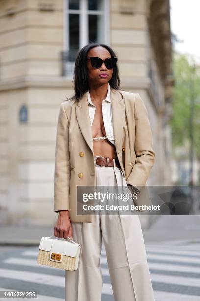 Emilie Joseph wears black large sunglasses, small earrings, a white linen V-neck / cropped laces shirt, a beige with gold buttons blazer jacket, a...