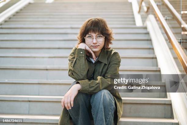 portrait of young teenage girl in eyeglasses sitting on stairs and looking at camera - cute college girl 個照片及圖片檔