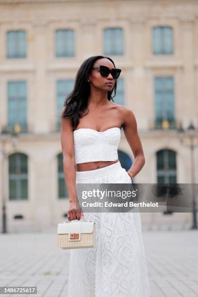 Emilie Joseph wears black large sunglasses, micro rhinestones earrings, a white embossed lace pattern heart shoulder-off / cropped top, a matching...