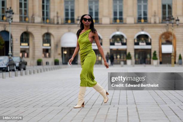 Emilie Joseph wears black large sunglasses, micro crystal earrings, a green satin / silk asymmetric one strap shoulder midi tube dress, gold...