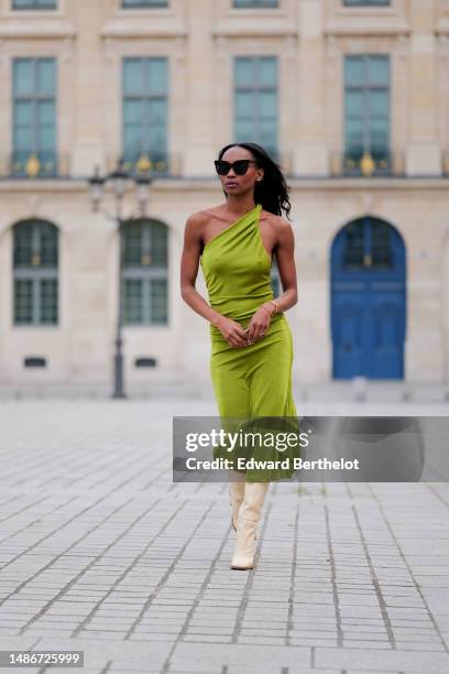 Emilie Joseph wears black large sunglasses, micro crystal earrings, a green satin / silk asymmetric one strap shoulder midi tube dress, gold...