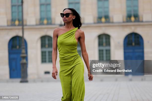 Emilie Joseph wears black large sunglasses, micro crystal earrings, a green satin / silk asymmetric one strap shoulder midi tube dress, gold...