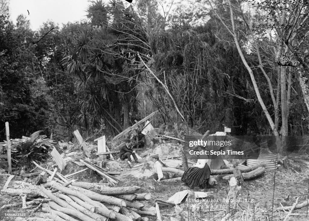 Ruins at Te Wairoa, near Rotorua, View of a man and two women sitting amongst timber, corrugated iron and logs at the site of a demolished buildings, against a backdrop of native bush. The remains of a watertank can be seen. on 10 Jan 1908