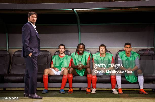 male soccer players on reserve sit on the bench beside their coach and watch the soccer game from tbe sidelines - shin guard stock pictures, royalty-free photos & images