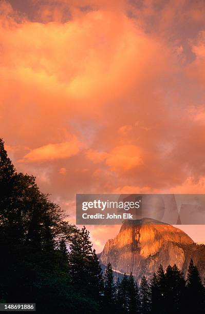 the clouds over the half dome are suffused with a brilliant orange glow, as the sun sets over the yosemite national park. seen from the sentinel bridge, which crosses the merced river - yosemite national park stock-fotos und bilder