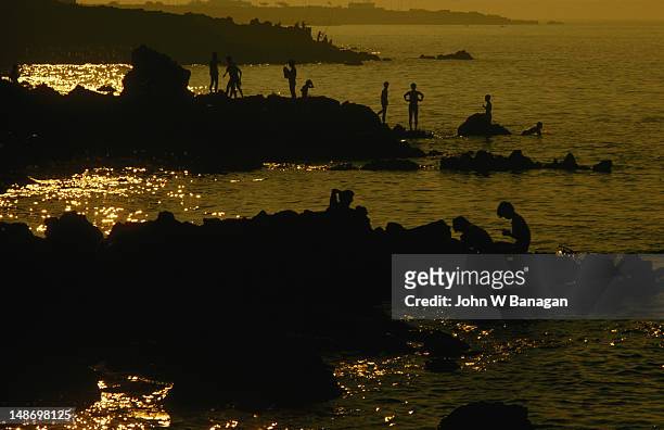 figures silhouetted against a sunset sky, playing by the water's edge on yongduam rock off the coast of jeju-si (cheju city) - jeju - fotografias e filmes do acervo