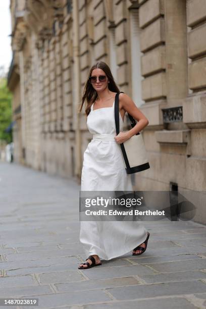 Diane Batoukina wears dark brown large squared sunglasses from Dior, a white square-neck / linen belted long dress, a pale gray fabric and black...