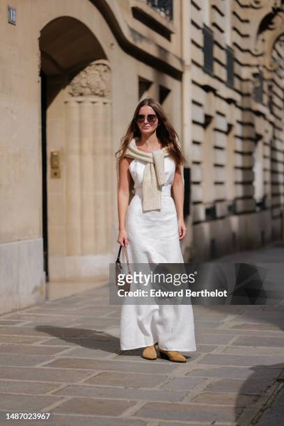Diane Batoukina wears dark brown large squared sunglasses from Dior, a white square-neck / linen belted long dress, a white latte ribbed wool...