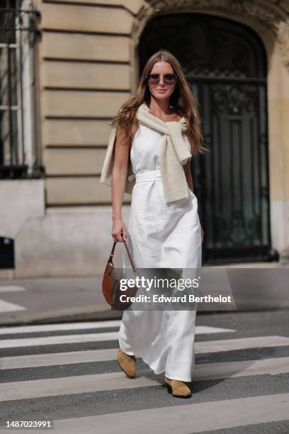 Diane Batoukina wears dark brown large squared sunglasses from Dior, a white square-neck / linen belted long dress, a white latte ribbed wool...