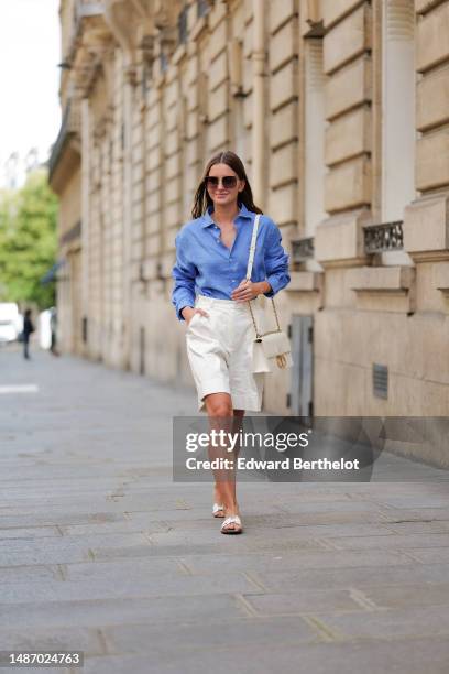 Diane Batoukina wears dark brown large squared sunglasses from Dior, a blue linen shirt from Figaret, white wide legs suit shorts, a gold Love ring...