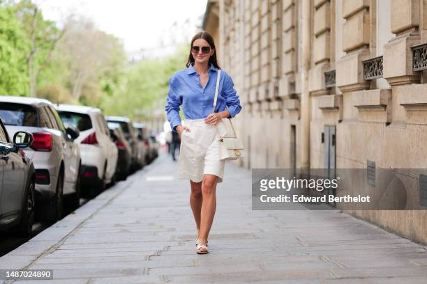 Diane Batoukina wears dark brown large squared sunglasses from Dior, a blue linen shirt from Figaret, white wide legs suit shorts, a gold Love ring...