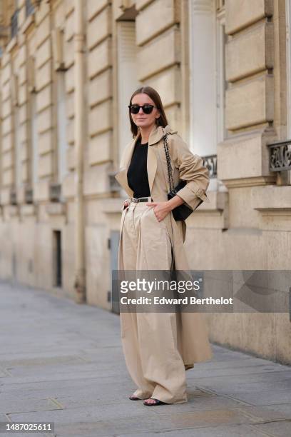 Diane Batoukina wears black circle sunglasses, a black ribbed halter-neck tank-top, a beige oversized trench coat from Filippa K, a black shiny...