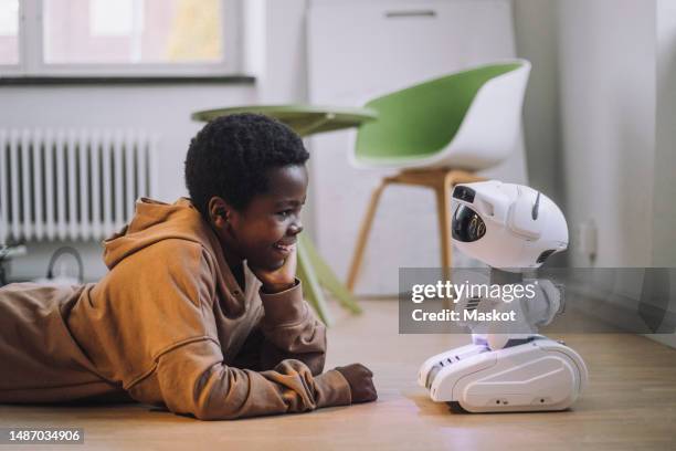 smiling boy talking with ai robot while lying on floor in innovation lab - prodotti infanzia foto e immagini stock