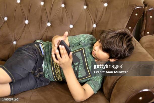 a young boy using mobile phone while lying on side on sofa in a living room at home. - pakistani boys stockfoto's en -beelden