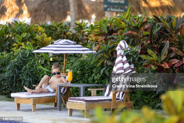 a little boy enjoys relaxing on a sun lounger under an umbrella by the pool - greater antilles stock pictures, royalty-free photos & images