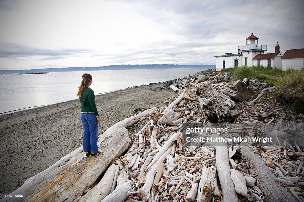 Woman standing on driftwood and looking towards Discovery Park Lighthouse.