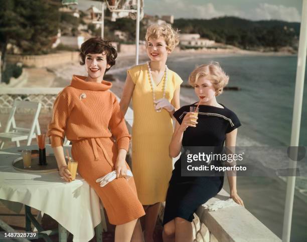Vacation scene of three female fashion models posed at a beachside cafe wearing, from left, orange, yellow and navy blue knitted rib dresses...