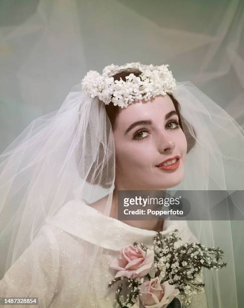 Posed studio portrait of a female fashion model in bridal wear, she wears an embroidered funnel necked bridal gown, veil and floral headwear, London,...