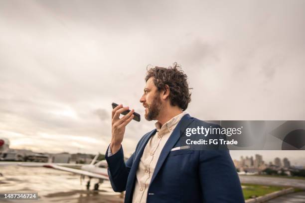 mid adult businessman sends a voice message while walking in a airport hangar - ditafone imagens e fotografias de stock