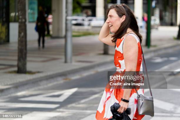 a lady walking down the street smiling and fixing her hair, wearing a white dress with orange flowers and a cute little black bag. - little black dress - fotografias e filmes do acervo