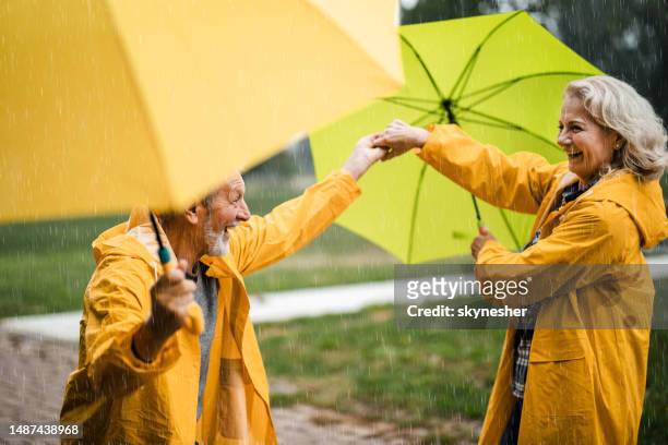 feliz pareja madura con impermeables amarillos divirtiéndose bajo la lluvia en la naturaleza. - young at heart fotografías e imágenes de stock