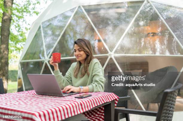 woman  sitting at table and using laptop in front of a geo dome glamping tent. - dome stock pictures, royalty-free photos & images