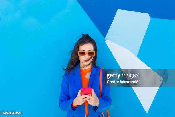 fashionable young woman using smart phone in front of blue wall - blue blazer stock pictures, royalty-free photos & images