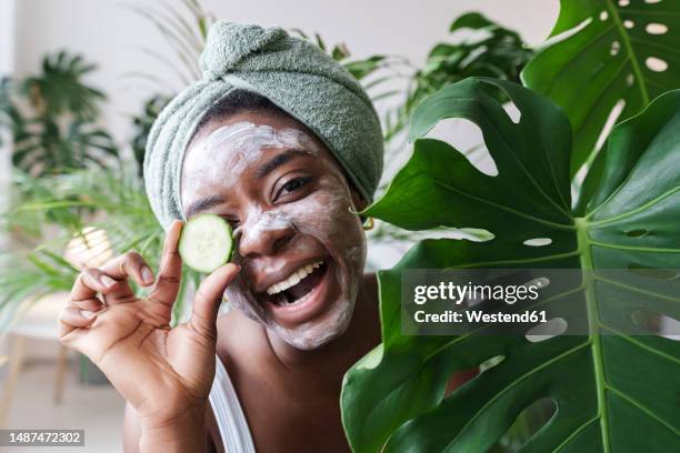 happy woman holding slice of cucumber next to monstera leaf - cucumber eye mask stock pictures, royalty-free photos & images