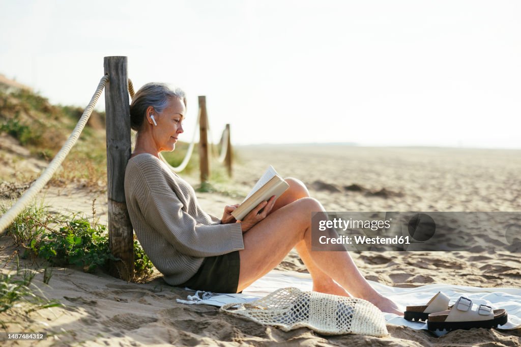 Mature woman wearing wireless in-ear headphones reading book at beach