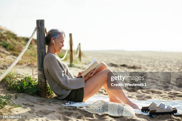 mature woman wearing wireless in-ear headphones reading book at beach - woman reading book stock pictures, royalty-free photos & images