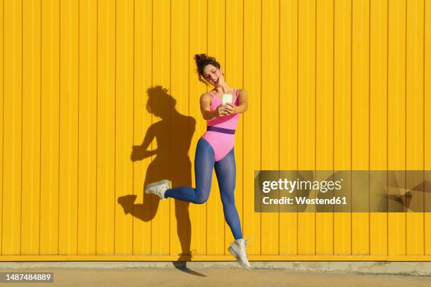 happy woman dancing and taking selfie through smart phone in front of yellow wall - human body foto e immagini stock