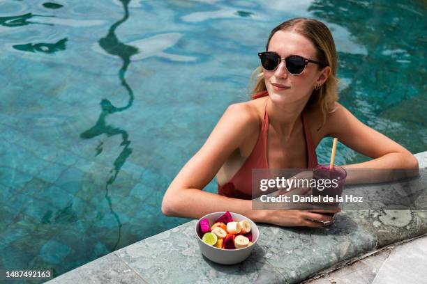 young blonde woman relaxing in the swimming pool drinking vegetable juice and eating healthy bowl of fruit. copy space. - tropical fruit imagens e fotografias de stock