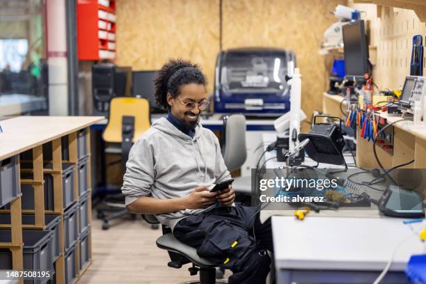 lächelnder elektroingenieur mit telefon, während er an der werkbank im maker-raum sitzt - stem cells human stock-fotos und bilder