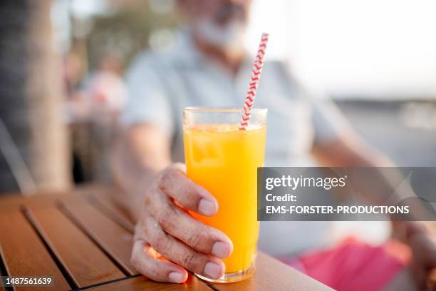 expatriate modern active senior man in his 60's enjoys time at the beach with healthy fresh orange juice - drinking straw photos et images de collection