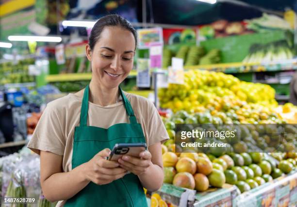 happy retail clerk using her phone while working at the supermarket - twitter the company of tweets stockfoto's en -beelden