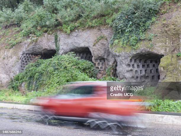 Car speeds along a stretch of the historic Via Cassia in Viterbo and on the tuffaceous part in the background we see ancient columbarium. Thus the...