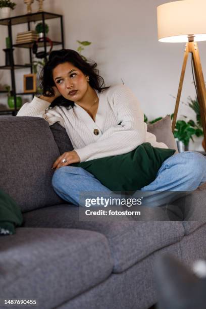 young woman sitting on the sofa, head in hand, contemplating life's problems - mood swings stock pictures, royalty-free photos & images