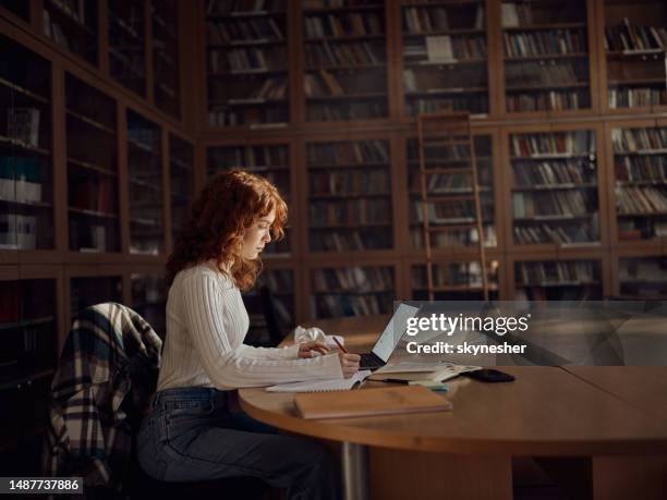 female student taking notes while e-learning in library. - biblioteca imagens e fotografias de stock