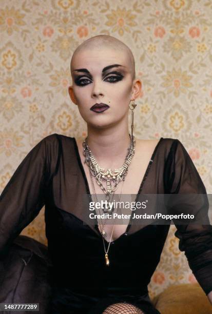 Portrait of a female punk, wearing goth style clothing, dark eye make-up and a shaved head, posed in a bedroom in London in 1983.