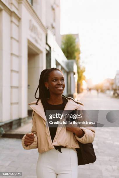 young black woman in light jacket and white jeans walking along city street - dark skin tone stock pictures, royalty-free photos & images