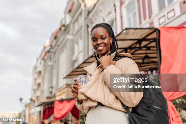 smiling black female traveler in light clothes looking at camera holding plastic coffee cup - cream coloured jacket stock pictures, royalty-free photos & images