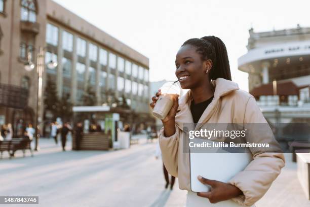 young black woman in beige jacket smiling walking around town square - ice coffee drink stock pictures, royalty-free photos & images