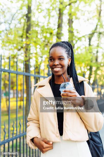 young black woman strolling by public park holding coffee cup and backpack - cream coloured jacket stock pictures, royalty-free photos & images