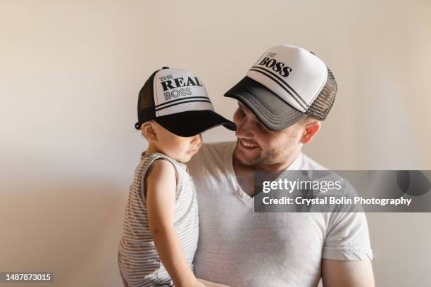 a 31-year-old father holding his 1-year-old son while they wear matching baseball caps for father's day that say "the boss" on dad's hat & "the real boss" on the baby boy's hat, neutral background with copy space mostly black & white colors - matching outfits stock pictures, royalty-free photos & images