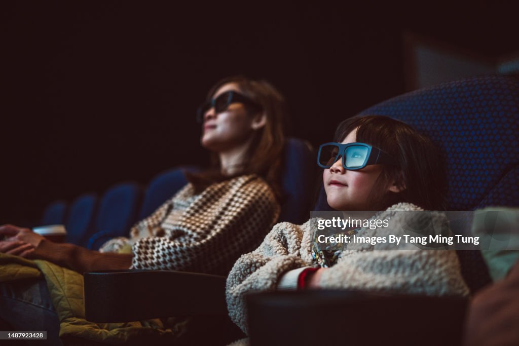 Mom & daughter watching movie with 3D glasses in cinema hall at movie theatre