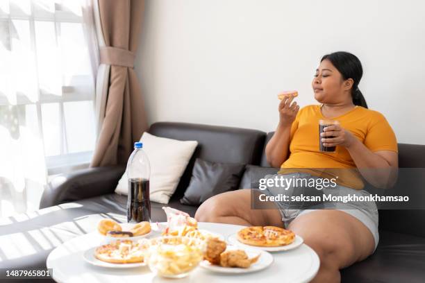 plus size woman sitting on sofa and eating junk food. - fat people eating donuts foto e immagini stock