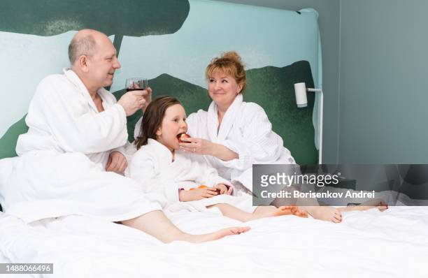a man, a woman with a daughter, a child of 7 years old, in dressing gowns, relax on a day off in the evening. they lie on the bed drinking wine and eating fruit. - lebanon wine stock pictures, royalty-free photos & images