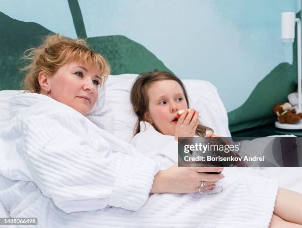 a woman with her daughter, a child of 7 years old, in dressing gowns, rest on the weekend in the evening. the family is resting on the bed watching tv, eating fruit, the woman is drinking red wine. - lebanon wine stock pictures, royalty-free photos & images