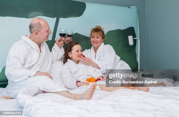 a man, a woman with a daughter, a child of 7 years old, in dressing gowns, relax on a day off in the evening. they lie on the bed drinking wine and eating fruit. - lebanon wine stock pictures, royalty-free photos & images