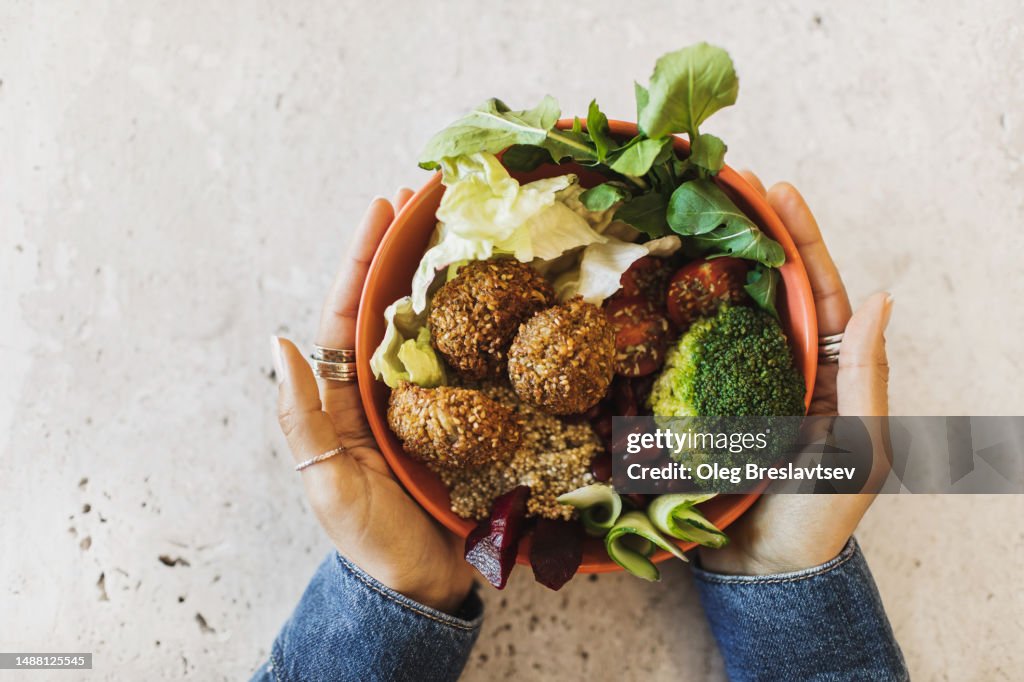 Woman's hands holding bowl with Falafel, broccoli, greens, cherry tomatoes, red beans, beetroot and quinoa